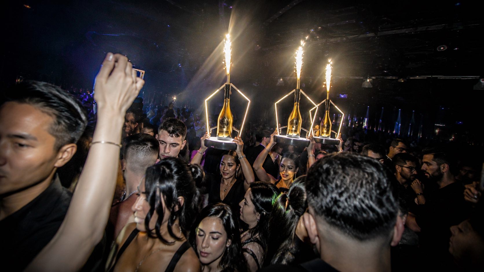 Bottle service carried through the crowd, sparklers lit, in a packed nightclub.